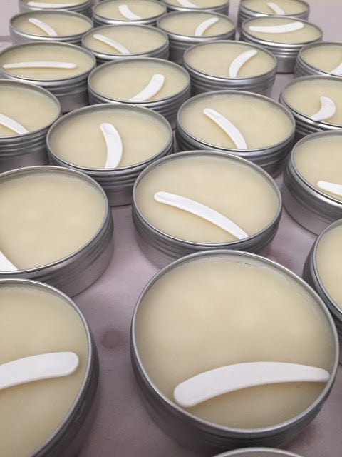 Multiple tins of balm with a white spatula on a purple background