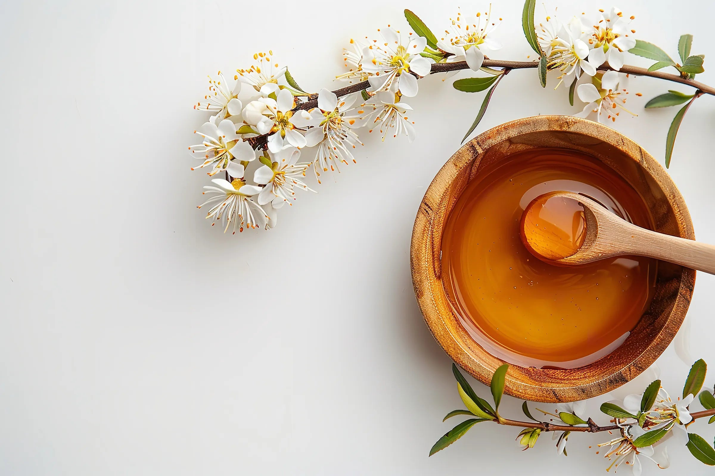 Wooden bowl of honey with a wooden spoon, surrounded by white flowers and green leaves on a light background