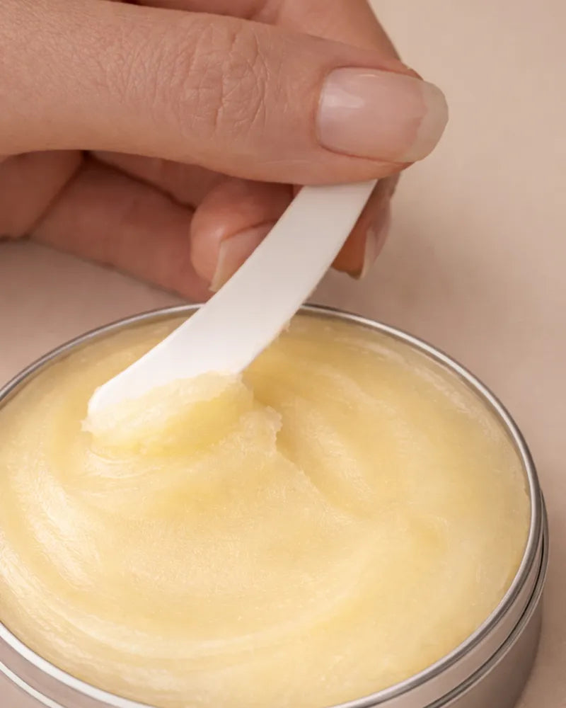 Hand using a white spatula to scoop out balm from a metal tin.